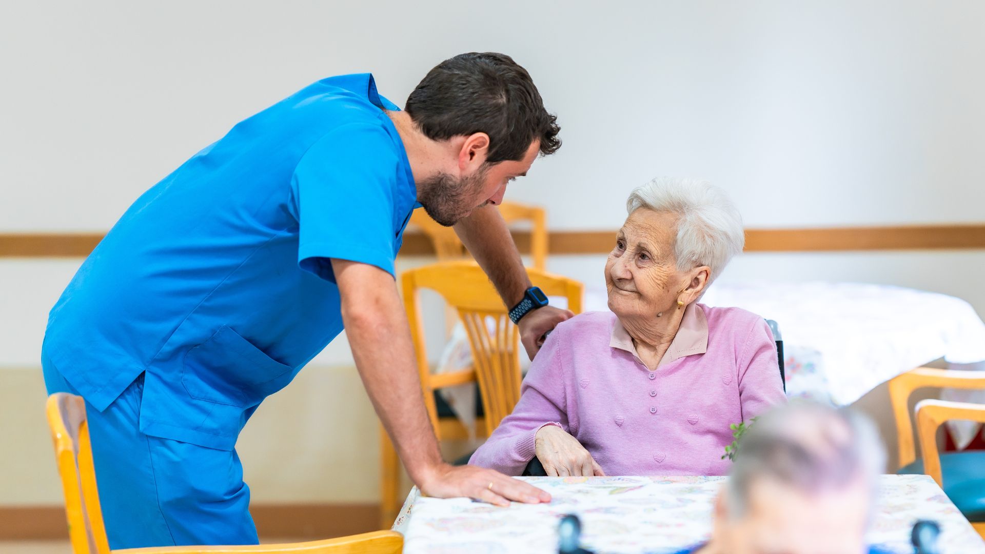 En vårdare står bredvid en äldre kvinna som sitter vid ett bord. Vårdaren lutar sig fram och samtalar med kvinnan i en ljus och trivsam miljö. På bordet finns en duk och några föremål, och i bakgrunden syns fler stolar och bord.